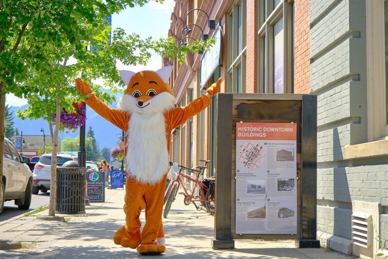 Fernie Fox Hotel Mascot walking down 2nd Ave next to a Discover Fernie kiosk.