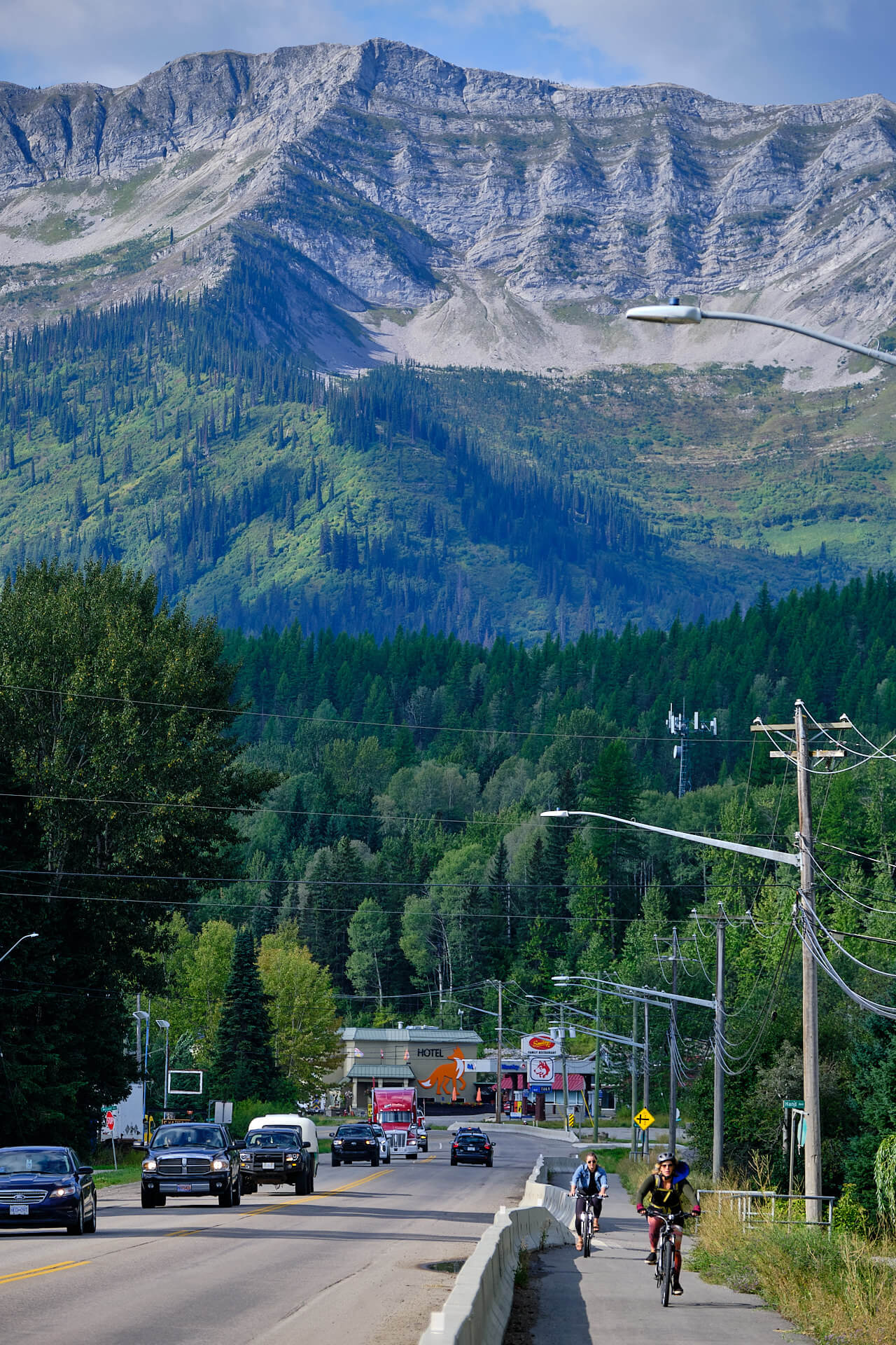 Fernie Fox Hotel along Hwy 3 in Fernie during the spring, the best time of year to visit Fernie. Cars driving down road, people cycling and walking along foot path.
